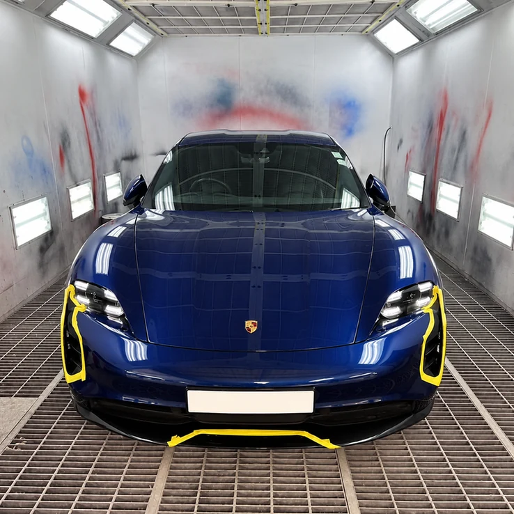 Blue Porsche inside a spray booth with yellow masking tape around the front bumper.