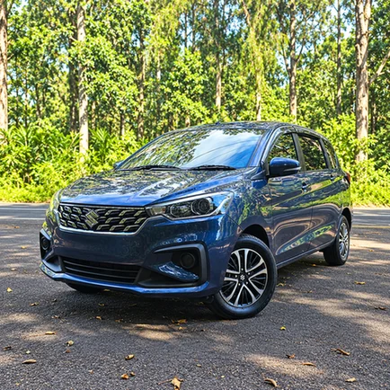 Blue Suzuki Ertiga parked beside a forest, viewed from the front and side.