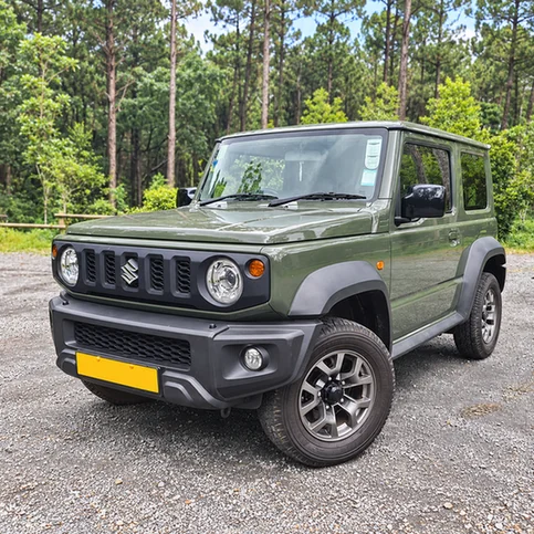 Olive green Suzuki Jimny parked on gravel with a forest behind it.