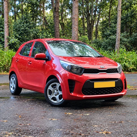 Red Kia Picanto parked on wet pavement beside a wooded road.