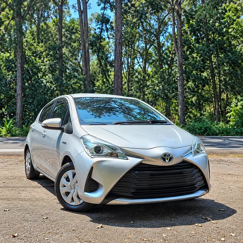 Silver Toyota Vitz Hybrid parked on a roadside with trees behind it.