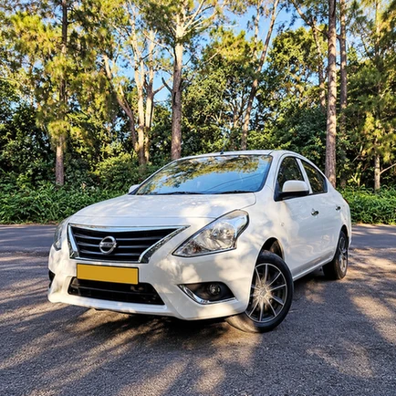 White Nissan Latio sedan parked beside a forest in dappled sunlight.