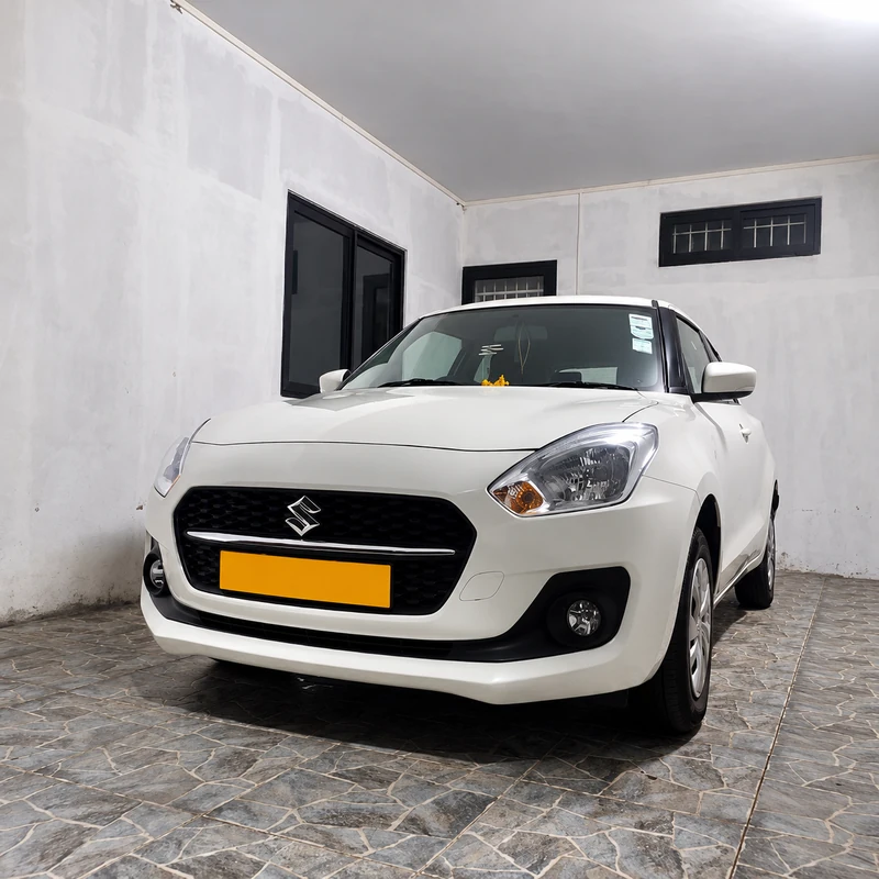 Front view of a white Suzuki Swift parked on paving beside tropical plants.